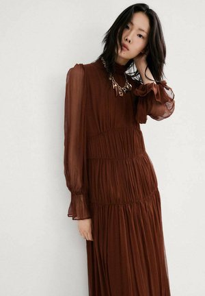 Młoda kobieta o czarnych włosach, ubrana w długą, brązową sukienkę z przezroczystymi rękawami oraz masywny złoty łańcuchowy naszyjnik, pozująca na tle jednolitego białego tła.