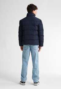 Giacca a vento blu navy con cuciture a canale, colletto alto e maniche lunghe. Indossata con jeans azzurri chiari e sneaker nere.