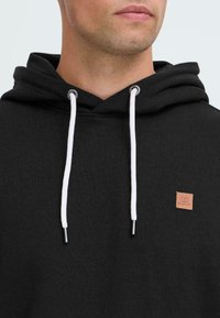 Uomo che indossa un hoodie nero con cordini bianchi e un piccolo patch quadrato marrone sul petto.