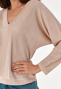 Pull en tricot beige clair avec décolleté en V, texture côtelée, coupe décontractée et manches longues. Présente un léger éclat et un design confortable.