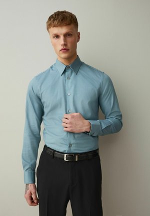 Ung mand iført en lyseblå skjorte med knapper og sorte bukser, justerer sin skjorte med venstre hånd mod en ensartet lys baggrund.