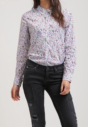 Vrouw draagt een wit shirt met lange mouwen met een klein bloemenprintje en zwarte, gescheurde skinny jeans.