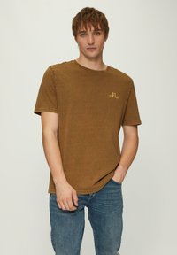 Bruin katoenen T-shirt met ronde hals, korte mouwen en een subtiele gele logo op de linkerkant van de borst. Gecombineerd met blauwe denim jeans.