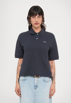 Kobieta o ciemnych włosach i kolczykach, ubrana w granatową koszulkę polo z napisem "CLOSED" oraz jasnoniebieską dżinsową spódnicę, stojąca na jednolitym tle.