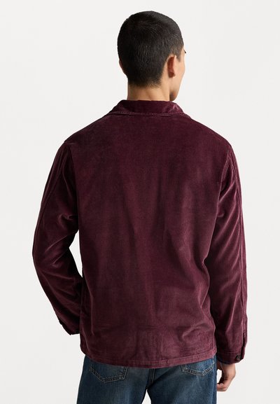 Burgundy corduroy jacket with long sleeves, textured stripes, and a collar. Features button cuffs and a straight hem, paired with blue jeans.