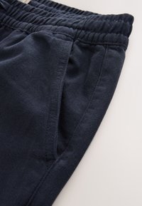 Tamsiai mėlynos laisvalaikio kelnės turi elastingą juosmenį ir įstrižas šonines kišenes, pagamintos iš minkštos, tekstūruotos medžiagos su matomomis siūlėmis.