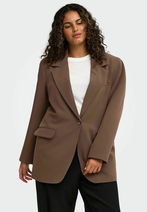 Femme aux cheveux bouclés portant un blazer marron oversize sur une chemise blanche et un pantalon noir, posant devant un fond clair uni.