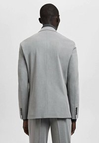 Man die van de camera af staat, gekleed in een lichtgrijs colbert en bijpassende broek tegen een effen witte achtergrond.