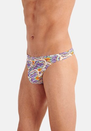 Männlicher Oberkörper, der bunte, abstrakt gemusterte Slips in Lila, Orange und Weiß trägt, steht vor einem einfarbigen Hintergrund.