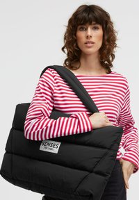 Schwarze Stepp-Shoppertasche mit gepolstertem Design und einem Schulterriemen, versehen mit einem sichtbaren weißen Logolabel. Die Tasche wird zu einem gestreiften Oberteil getragen.