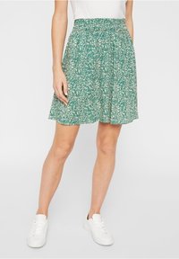 Zelená tištěná sukně s květinovým vzorem, se shromážděným pasem, délka ke kolenům a splývavý materiál. Nosič nosí s bílými teniskami.