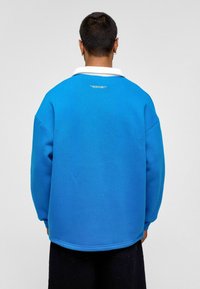 Blauwe sweatshirt met een witte kraag, zachte textuur,Relaxed fit en gestikte details op de rug, gedragen met donkere broek.