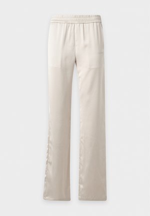 Jeune femme en débardeur court blanc, pantalon ample crème, baskets noires et blanches, tenant un petit sac à main noir sous le bras.