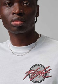 Primer plano de una persona que lleva una camiseta blanca de Air Jordan con texto rojo "Flight" y un collar de cadena plateada sobre un fondo gris.