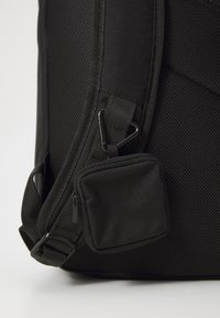 Sac à dos noir en matériau en maille, avec des bretelles ajustables, une petite poche zippée et des accents en métal pour attache.