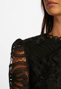Top en dentelle noire avec une broderie florale délicate, des manches à motifs transparentes et un col festonné avec des découpes.