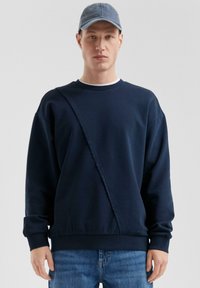 Felpa blu navy con una striscia diagonale testurizzata, colletto a girocollo e maniche lunghe. Abbinata a un cappellino in denim azzurro e jeans.