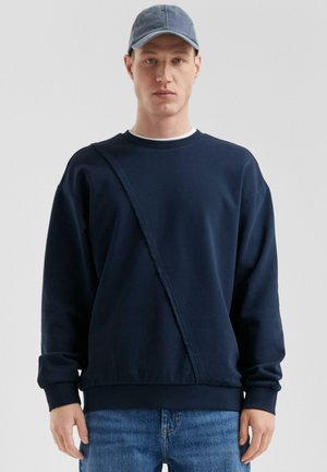 Marineblauwe sweatshirt met een diagonale gestructureerde streep, ronde hals en lange mouwen. Gekleed met een lichtblauwe denim pet en jeans.