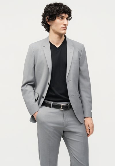 Νέος άνδρας με σγουρά μαλλιά που φοράει ανοιχτό γκρι κοστούμι, μαύρο μπλουζάκι με V λαιμόκοψη και μαύρη ζώνη, κοιτάζοντας στο πλάι με το ένα χέρι στην τσέπη.