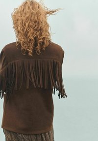 Brown suede jacket featuring long fringes across the shoulders, with a textured surface and a relaxed silhouette. Background is light blue.