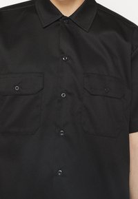 Chemise noire à manches courtes avec un col boutonné, deux poches plaquées et des boutons en métal noir, fabriquée dans un tissu lisse à texture mate.