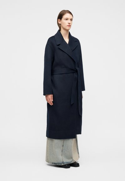 Γυναίκα που φοράει μακρύ σκούρο μπλε παλτό με ζώνη πάνω από ένα ανοιχτό μπλε τζιν φούστα και μαύρα παπούτσια, στέκεται μπροστά σε λευκό απλό φόντο.