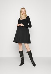 Vestido preto de mangas longas com decote quadrado e saia rodada, feito de tecido texturizado. Combinado com botas de couro pretas até ao joelho.
