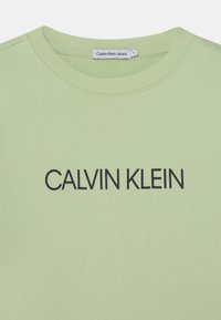 Světl zelený svetr s kulatým výstřihem, na přední straně s nápisem "CALVIN KLEIN" v tučných černých písmenách. Měkká textura.