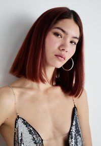 Top in paillettes nere e argento con spalline sottili e scollatura profonda, caratterizzato da un dettaglio in catena metallica. Texture liscia. Capelli corti di colore rosso.