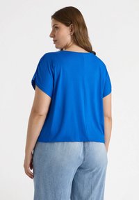 Blaues kurzärmliges Oberteil mit einem lockeren Schnitt. Auffällige Nahtdetails auf der Rückseite, kombiniert mit hellblauen Hochtaillenhosen. Sanfte Textur.