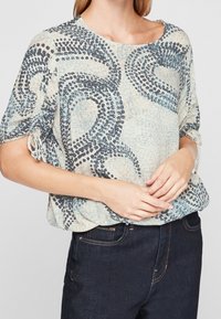 Blusa leggera di colore crema con motivi astratti in blu navy, con maniche corte e dettagli arricciati ai polsini. Abbinata a jeans scuri.