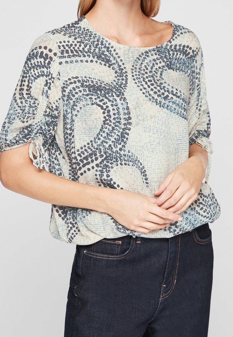 Blusa leggera di colore crema con motivi astratti in blu navy, con maniche corte e dettagli arricciati ai polsini. Abbinata a jeans scuri.