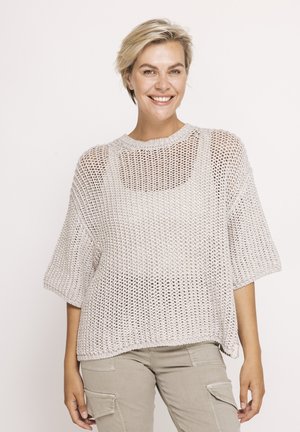 Smiling woman with short blonde hair wears loose, light gray knit sweater over beige cargo pants, posing against plain light background.