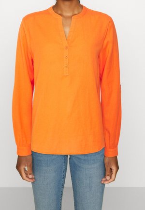 Chemise orange à manches longues avec col mao, trois boutons au niveau du cou et poignets élastiqués, associée à un jean bleu.