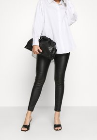 Svarta leggings i konstläder, matchade med en vit skjorta med knappar. En texturerad svart clutchväska med snörning. Svarta öppna tofflor.