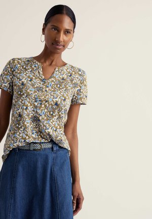 Kvinde med guldringe i ørerne iført en blomstret kortærmet top, stoppet ned i en blå denimnederdel med flettet bælte, stående foran en lys baggrund.