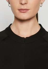 Černý top s kulatým výstřihem a krátkým zipem vpředu. Tkanina se zdá být hladká a mírně elastická.