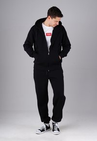 Man wearing black hoodie, black sweatpants, black and white sneakers, and white T-shirt with red text looking downward, hands in pockets, gray background.
