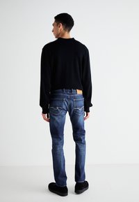 Suéter negro de mangas largas combinado con jeans azul desgastado. Los jeans tienen bolsillos traseros con detalles de costura y una etiqueta de marca de cuero. Zapatos negros.