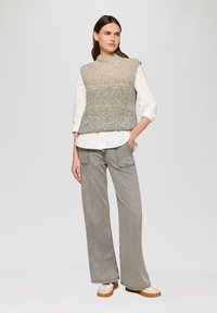 Lichtgrijze, gebreide mouwloze top met een hoge hals, gedragen over een witte button-up blouse, gecombineerd met wijde grijze jeans en beige sneakers.
