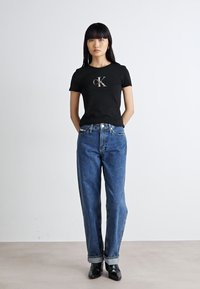 Melnas kokvilnas T-krekls ar sudraba "cK" logotipu, apvienots ar augstās jostasvietas zilajiem džinsiem, kas salocīti pie potītēm, valkāts ar melnām kurpēm.