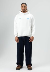 Witte hoodie met voorzak, voorzien van blauwe tekstaccenten. Gecombineerd met donkerblauwe wijde jeans en bruine veterlaarzen.