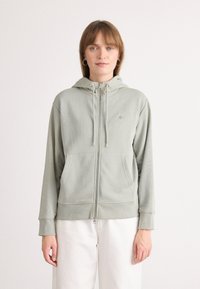 Felpa con zip verde chiaro realizzata in tessuto morbido, dotata di cappuccio, tasche frontali, polsini a coste e un piccolo dettaglio con logo.