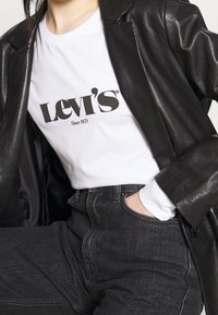 Weißes Levi's T-Shirt mit schwarzem Logo, kombiniert mit dunkelgrauen Jeans und einem schwarzen Lederblazer, der sichtbare Nähte und Taschen aufweist.
