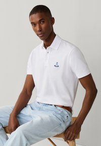 Man met een wit poloshirt met een klein blauw embleem, lichtblauwe spijkerbroek en een bruine riem, zittend op een houten kruk tegen een effen achtergrond.