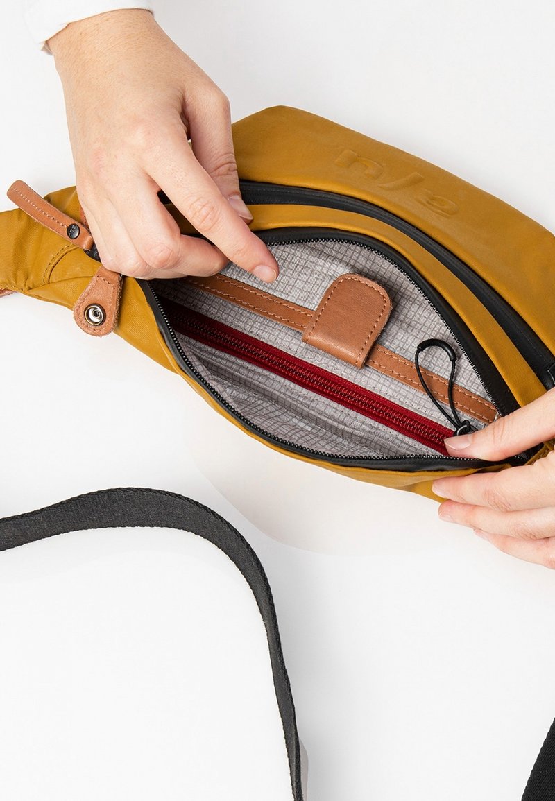 Des mains ouvrent un sac jaune moutarde avec une fermeture éclair noire, montrant ses compartiments intérieurs beige et rouge ainsi qu’une sangle noire réglable.