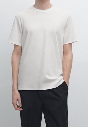 Person iført en almindelig hvid T-shirt med korte ærmer og sorte bukser, den ene hånd i lommen, vist fra nakken til hofterne mod en ensfarvet baggrund.