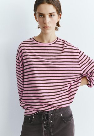 Mladá osoba s krátkymi vlasmi, oblečená v ružovo-hnedom pruhovanom tričku s dlhým rukávom a čiernych šnurovacích nohaviciach, stojaca na neutrálnom pozadí.