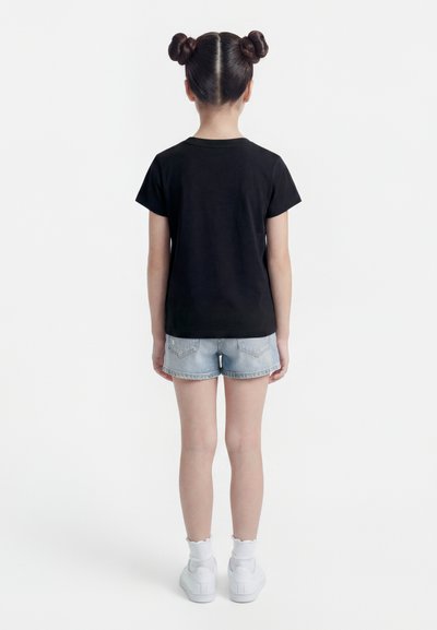 Παιδί που στέκεται πλάτη φορώντας μαύρο μπλουζάκι, ανοιχτόχρωμα denim σορτς, άσπρες κάλτσες και άσπρα αθλητικά παπούτσια, με μαλλιά χτενισμένα σε δύο κότσους.