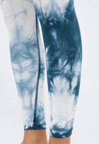 Κάτω άκρα που φορούν μπλε και λευκά κολάν με σχέδιο tie-dye και εμφανείς μαύρες ραφές στο πλάι.